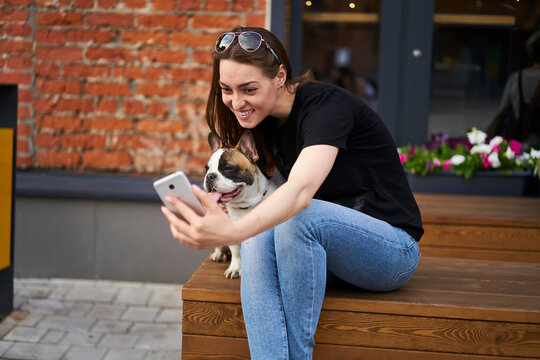 Happy hipster girl clicking selfie photo on modern smartphone web camera sitting on bench with french bulldog, cheerful woman posing with doggie friend and taking images for own blog about pet