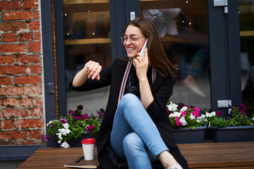 Happy hipster girl in classic spectacles smiling during positive cell talking in roaming, joyful...