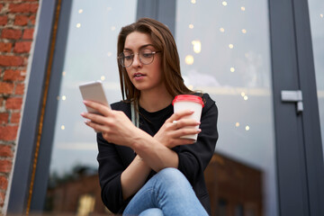 Fototapeta premium Young hipster girl in optical eyeglasses holding takeaway coffee to go while resting outdoors and using cellphone for online messaging, millennial woman reading web content while making booking