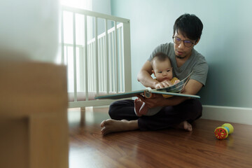 Asian Father reading book or bedtime stories to his little cute baby son while sitting on the floor...
