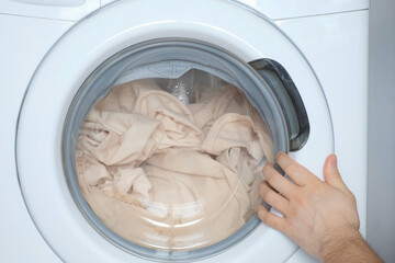 Man's hands loading the washing machine with beige bed linen, laundry at home. Man is doing his laundry and closing the door of washing machine. Everyday life, housework of young man.