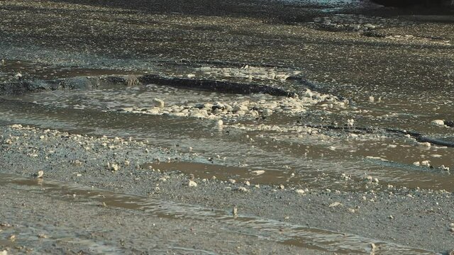 Close-up cars drive along a bad road with holes, muddy puddles and cracks. A crumbling road with potholes. Old asphalt pavement made in violation of the technology of work. Taxpayer money is wasted.