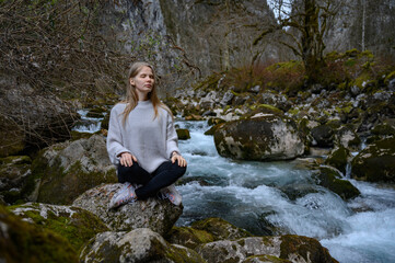 Girl sits on a large stone and meditates near a waterfall next to a mountain river.
