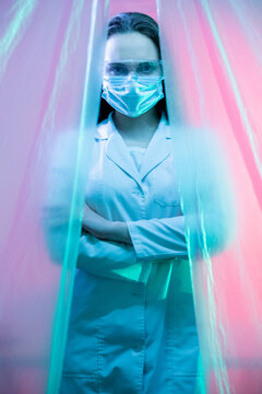 Scientist Portrait. Biochemical Research. Microbiology Study. Confident Female Lab Technician With Crossed Arms In Face Mask In Pink Blue Neon Light Isolated Behind Plastic Drapes.