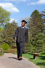 Handsome young black college graduate wearing cap and gown walks proudly along a path