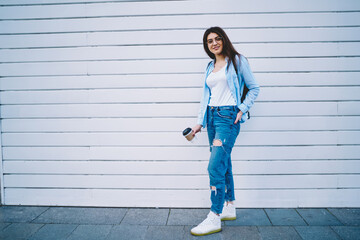 Full length portrait of stylish Caucasian woman 20 years old dressed in casual wear posing outdoors, pretty hipster girl holding takeaway coffee to go in hand smiling at camera at urban setting