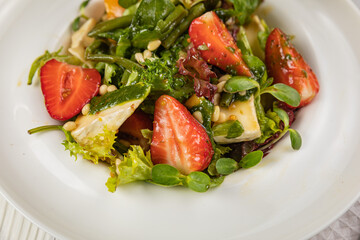 Salad with strawberries, brie cheese, tomatoes and greens. On a white plate and white table.

