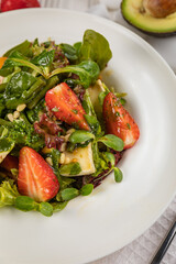Salad with strawberries, brie cheese, tomatoes and greens. On a white plate and white table.
