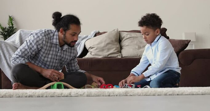 African father and son having fun play toy rail road seated on carpet in living room. Family spend weekend together at home enjoy playtime use modern cool plaything. Free time, leisure, hobby concept