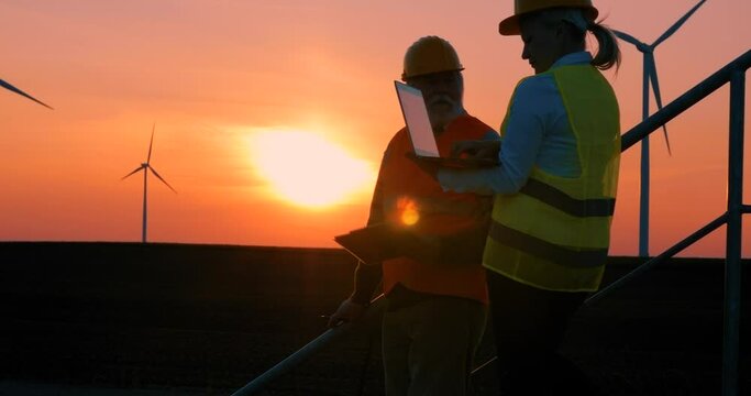 Old man engineer and a female worker descend the stairs from a windmill at sunset