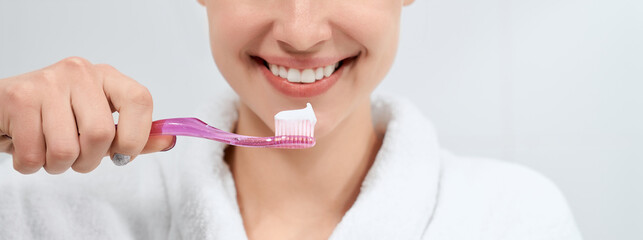 Close up of smiling young woman in white robe holding toothbrush with paste on light background. Concept of preparing for cleaning teeth at home with good mood.
