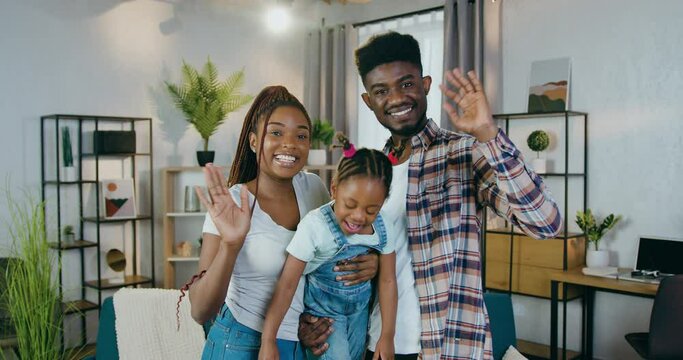 Portrait of young african american family smiling and waving hands on camera while standing at modern living room. Happy parents with cute daughter enjoying time spending together.