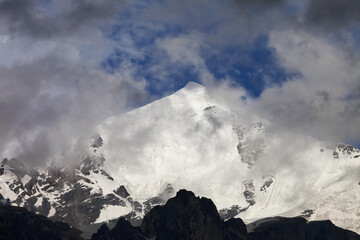 High snowy mountains with glacier, rocks and blue sky with dark fog before storm