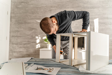 Obraz premium One Man with beard alone Putting Together Self Assembly Furniture at Home holding electric screwdriver screwing and putting together pieces - half length front view DIY concept real people copy space