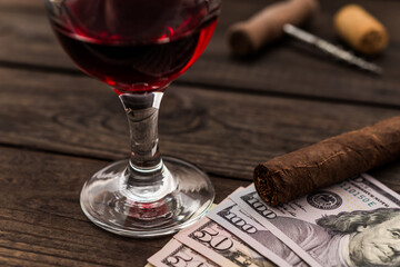 Glass of red wine with money and cuban cigar with corkscrew and corks on an old wooden table. Close up view, focus on the cuban cigar
