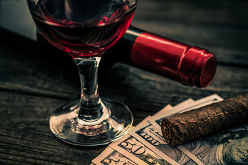 Bottle of red wine with a glass of red wine and cuban cigar with lot of money on an old wooden table. Close up view, focus on the cuban cigar