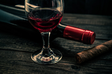Bottle of red wine with a glass of red wine and cuban cigar on an old wooden table. Angle view, focus on the cuban cigar