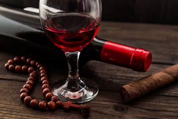 Bottle of red wine with a glass of red wine and cuban cigar with rosary on an old wooden table. Angle view, focus on the cuban cigar