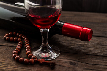 Bottle of red wine with a glass of red wine and rosary on an old wooden table. Angle view, focus on the glass of red wine