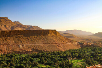 View over green palm tree oasis on dry arid brown mountain plateau with road in morning twilight - Tinghir, Morocco