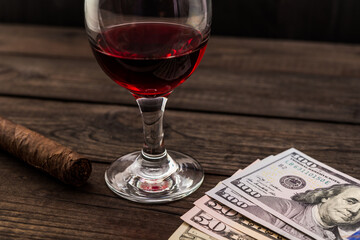 Glass of red wine and money with cuban cigar on an old wooden table. Angle view, focus on the money