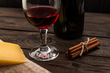 Bottle of red wine with a glass of red wine and a piece of parmesan and cinnamon sticks on an old wooden table. Angle view, focus on the glass of red wine