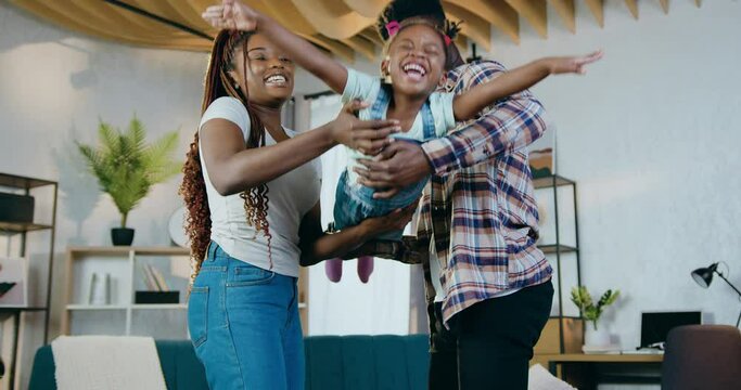 Cheerful african man and woman waving on hands their cute little daughter while staying at home. Young family in casual clothes spending free time actively.