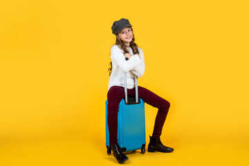 cheerful child in headwear with valise for travelling on yellow background, vacation