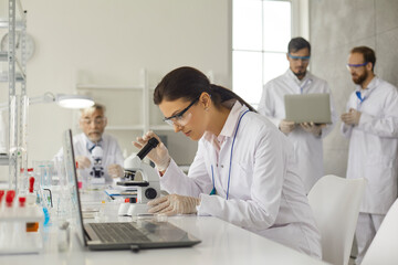 Fototapeta premium Virologist doing research studying in medical laboratory. Concentrated female virologist in goggles and latex gloves sits at table in laboratory and examines cells under microscope.