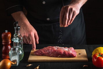 Professional chef sprinkles the raw beef meat with salt. Preparing meat before baking. Working environment in kitchen of restaurant or cafe