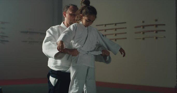 Cinematic shot of budokai fighter master helping to tie belt of kimono of female pupil before Aikido training in martial arts school. Concept: learning, sports, recreation, defence, religious beliefs.