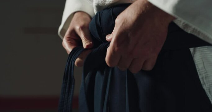 Cinematic macro of mature female budokai fighter tying belt of kimono before practicing Aikido in martial arts school. Concept: healthy lifestyle, sports, recreation, defense, religious beliefs