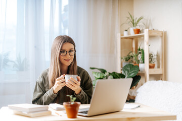 Thinking Freelance woman in glasses with coffee cup, notebook and laptop working from home office with plants. Happy girl on workplace at the desk. Distance learning online education and work.