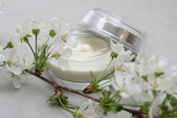 Fototapeta na wymiar Beauty cream in a glass jar on a light gray background. Decorated with white spring flowers. Unbranded skincare product. Cosmetic cream. Close up, selective focus.