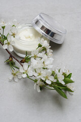 Fototapeta na wymiar Beauty cream in a glass jar on a light gray background. Decorated with white spring flowers. Unbranded skincare product. Cosmetic cream. Close up, selective focus, high angle. .