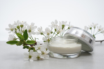 Obraz na płótnie Canvas .Beauty cream in a glass jar on a light gray background. Decorated with white spring flowers. Unbranded skincare product. Cosmetic cream. Close up, selective focus, side view.