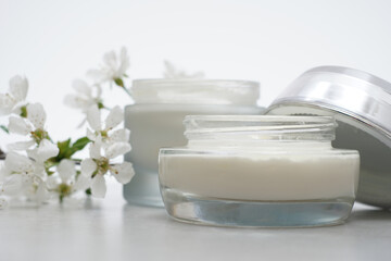 Beauty cream in glass jars on a light gray background. Decorated with white spring flowers. Unbranded skincare product. Cosmetic cream. Close up, selective focus, side view. - 433138711