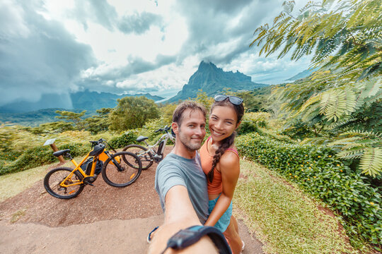 E-bike biking couple taking seflie on bikepacking travel vacation. Interracial tourists Asian woman, Caucasian man happy having fun on Moorea island ride, Tahiti, French Polynesia holiday adventure.