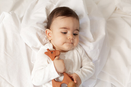 cute little mestizo girl lies in bed in a white bodysuit with cotton bedding, holding a wooden toy in her hands. High quality photo