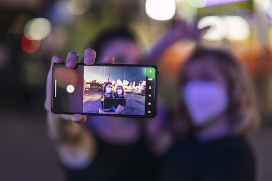 Cheerful Caucasian Female Friends With Masks Taking Photos At An Amusement Park In The Evening