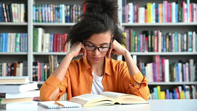 Tired exhausted African American female student sits at a table with books in the library, closed her eyes, massaging the bridge of her nose, headache and stress from overwork from learning, need rest