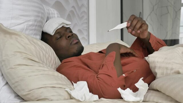 Sad African-American man with patch on head checks temperature suffering from coronavirus disease lying on bed at home closeup