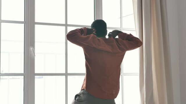Young African-American man in pajamas stretches body coming to large French window in early morning slow motion backside view