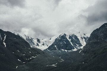 Dark atmospheric mountain landscape with glacier on black rocks in gray cloudy sky. Snowy mountains in low clouds in rainy weather. Gloomy mountain landscape with black rocky mountains with snow.