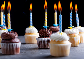 Close up side view image of an assortment of chocolate and vanilla cupcakes with white and brown frostings on. Each have a candle lit against dark background. Celebration, party, birthday concepts.