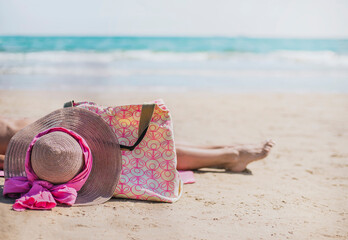Fototapeta na wymiar a beach bag next to a hat with a pink bow in the foreground. behind with selective blur, a woman lies sunbathing on a turquoise background of the mediterranean sea. vacation concept.