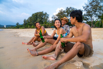 Fototapeta premium Group of Happy Asian man and woman friends sitting on the beach enjoy drinking beer with talking together at summer sunset. Male and female friendship relax and having fun outdoor lifestyle activity.