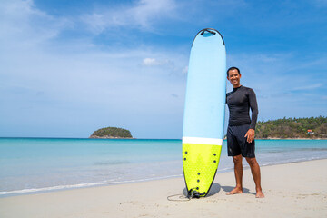 Attractive Asian man in wetsuit holding surfboard standing on tropical beach at summer sunny day....
