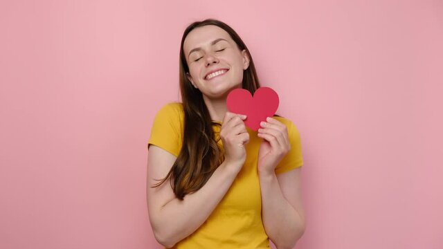Portrait of charming pretty young caucasian woman hold and pointing on small red heart, friendly happy looking at camera, wears yellow t-shirt, standing over pink studio background. Holiday concept