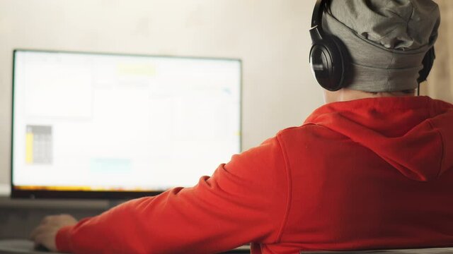 A programmer in a gray hat works in a home office in the evening sitting at a monitor screen, develops sites, copy space